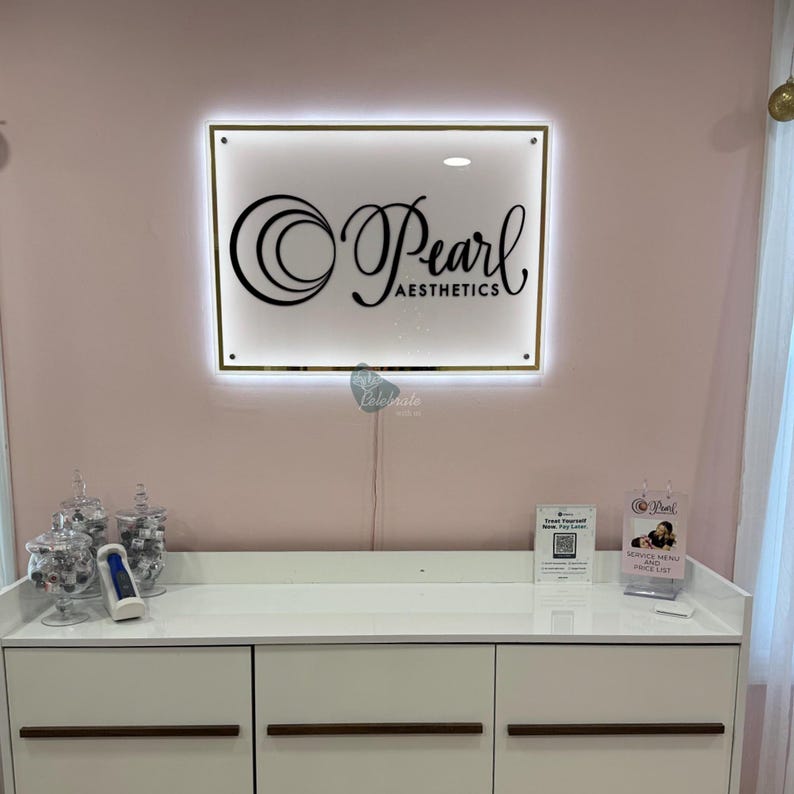 May include: A lit sign with the words "Pearl Aesthetics" in a cursive font hangs on a pink wall. Below the sign is a white cabinet with three drawers. Glass jars and a small device sit on the cabinet.