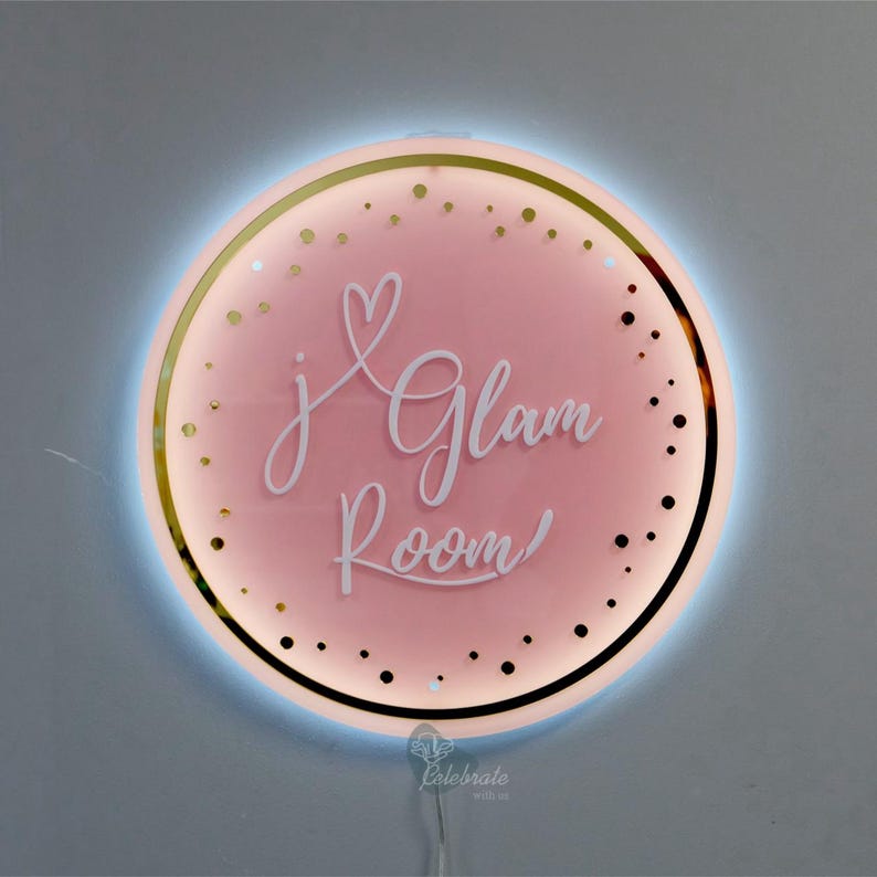 May include: A circular, pink illuminated sign with the words "j Glam Room" in a cursive font. The sign has a gold border and is decorated with gold dots. A blue light surrounds the sign, and a white cable extends from the bottom.