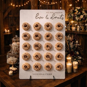 May include: A white sign with the text "All you need is love & donuts" displays rows of donuts. The donuts are light brown with chocolate sprinkles and nuts. The sign is on a wooden table, with candles and floral arrangements in the background.