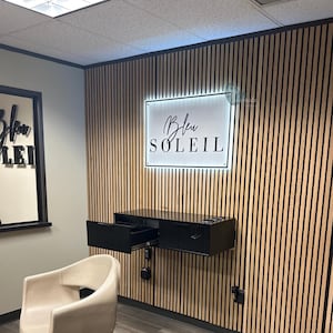 May include: Interior shot of a salon with a wooden slatted wall. A black floating cabinet with an open drawer is mounted on the wall. A sign with the text "Bleu Soleil" is illuminated above the cabinet. A cream-colored chair is in the foreground.