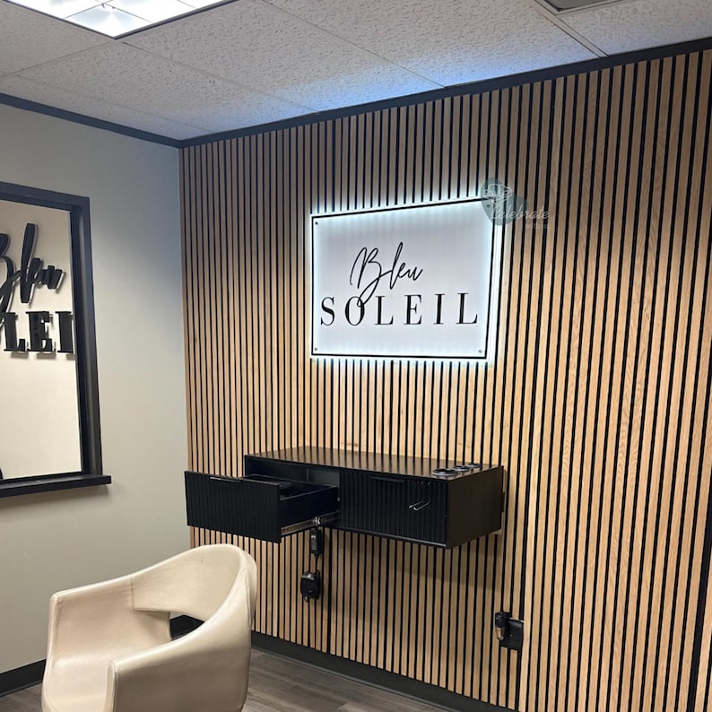 May include: Interior shot of a salon with a wooden slatted wall. A black floating cabinet with an open drawer is mounted on the wall. A sign with the text "Bleu Soleil" is illuminated above the cabinet. A cream-colored chair is in the foreground.