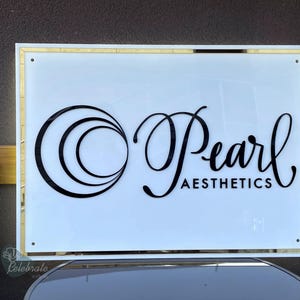 May include: A rectangular sign with a white background and gold trim. The sign features the words "Pearl Aesthetics" in black script and a stylized circular design. The sign is mounted on a dark surface.