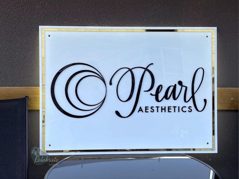 May include: A rectangular sign with a white background and gold trim. The sign features the words "Pearl Aesthetics" in black script and a stylized circular design. The sign is mounted on a dark surface.