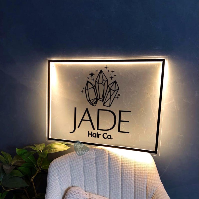 May include: Illuminated sign for "Jade Hair Co." featuring a black border and a frosted glass effect. The sign displays a crystal graphic above the business name. The sign is mounted on a dark blue wall.