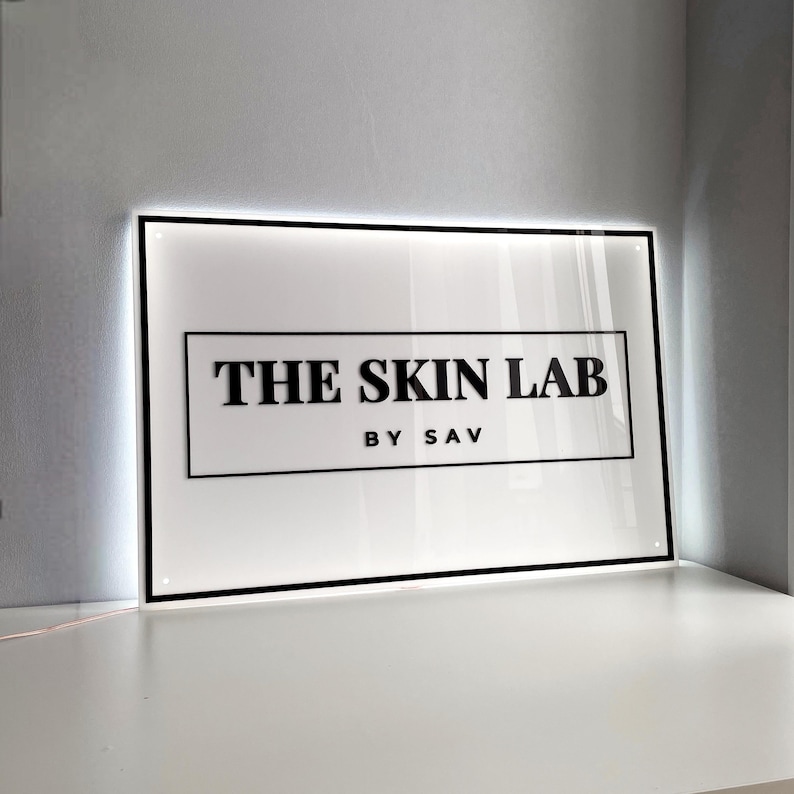 May include: A rectangular sign with the text "THE SKIN LAB" in bold black letters, with "BY SAV" below. The sign is white with a black border and is illuminated with a soft white light.