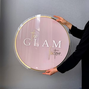 Puede incluir: Un letrero redondo de color rosa con un borde dorado y el texto "The Glam Factor". El letrero está siendo sostenido por la mano de una persona.