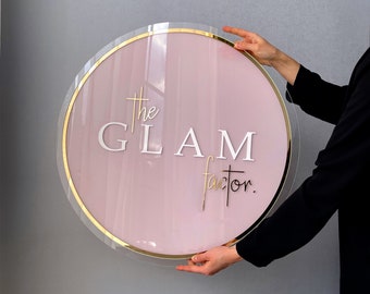 May include: A round, pink sign with a gold trim and the text "The Glam Factor." The sign is being held by a person's hand.