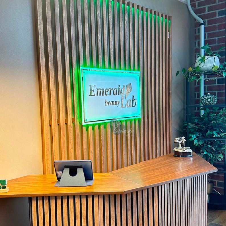 May include: A reception desk with a wooden countertop and a sign that reads "Emerald beauty Lab." The sign is illuminated with green light. A tablet sits on the desk, and the background features wooden slats.