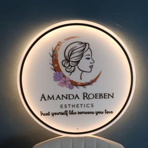 May include: A circular, illuminated sign with a white face and black edging. The sign displays a woman's profile, a crescent moon, and floral details. The text reads "AMANDA ROEBEN ESTHETICS" and "Treat yourself like someone you love."