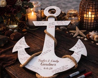 May include: A white wooden anchor-shaped guest book with rope detailing. The anchor is inscribed with the names "Taylor & Emily Gardner" and the date "6.6.26". Guests have written messages on the anchor. Two Sharpie markers are next to the anchor.