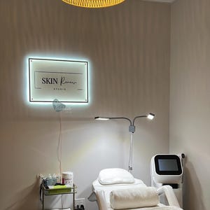 May include: A well-lit interior of a skin care studio. A sign with the text "SKIN Renew STUDIO" is mounted on the wall. A treatment bed, medical equipment, and a small metal cart with supplies are visible. A modern, minimalist aesthetic.