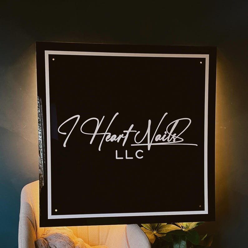 May include: A black square sign with a white border and the text "I Heart Nails LLC" in a script font. The sign is illuminated from behind, creating a warm glow.