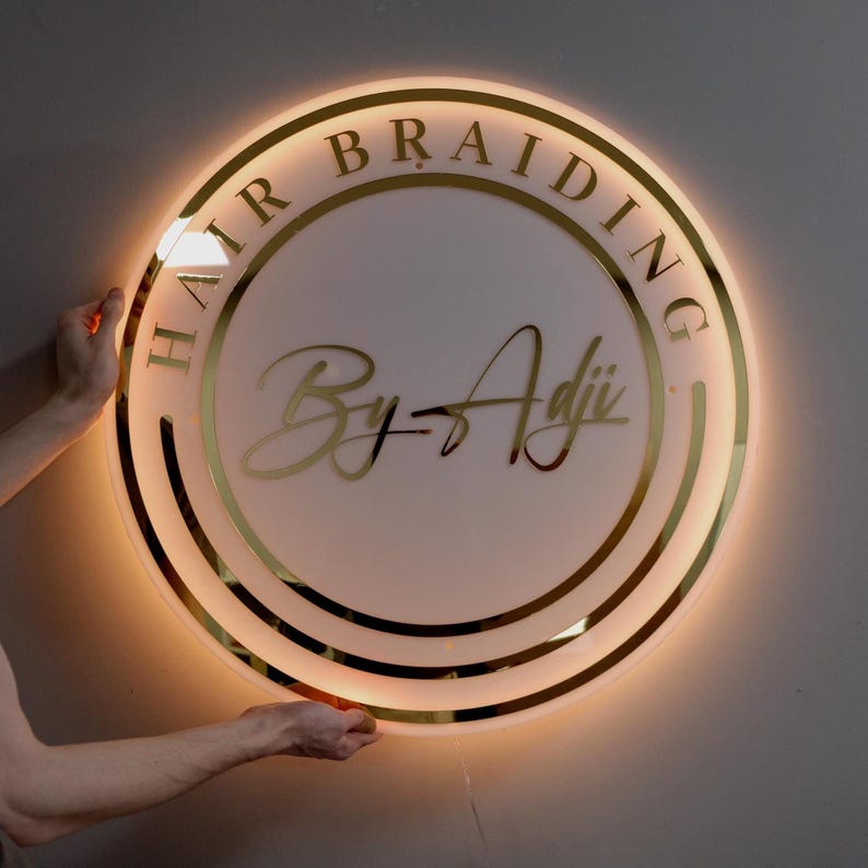 May include: A circular illuminated sign with gold lettering that reads "HAIR BRAIDING" around the edge. The centre features the text "By-Adji" in a stylish script. The sign emits a warm, golden light.