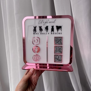 Puede incluir: Un letrero de acrílico rosa con fondo blanco, con el texto "Let's get social" y siluetas de perros. Incluye iconos de Instagram, Facebook y globo, junto con tres códigos QR. El letrero está diseñado para MAS Bully Rescue.