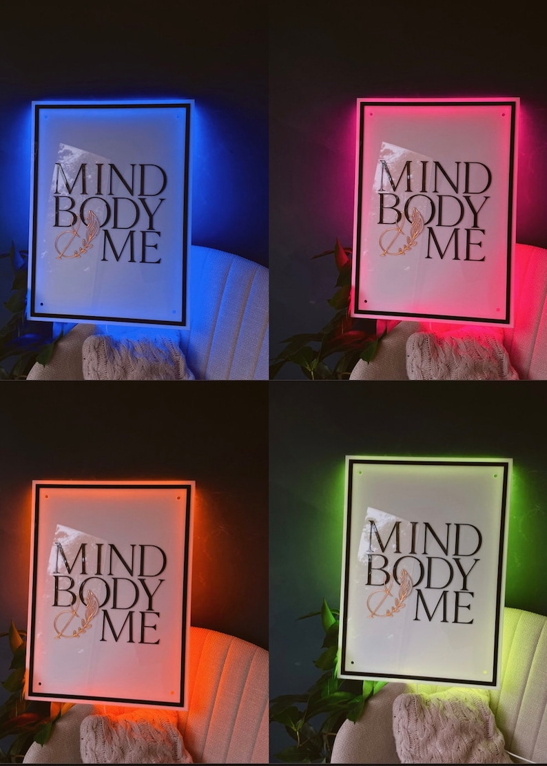 May include: A framed wall art piece with the text "MIND BODY ME" in black lettering, accented with a gold leaf design. The frame has a black border and is illuminated with a colored LED light. The image shows the art with blue, red, orange, and green lighting.