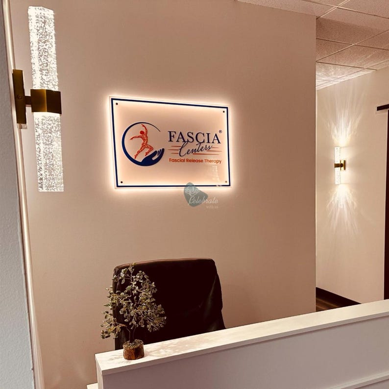 May include: An illuminated sign for Fascia Centers, featuring a logo with a stylized figure and the text "Fascia Centers, Fascial Release Therapy." The sign is mounted on a wall, with a reception desk and decorative lighting visible.