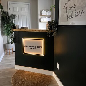 May include: Interior shot of a beauty salon reception area. The black reception desk features a glowing sign that reads "VEIL BEAUTY LOFT SKIN & BEAUTY." A round, woven rug sits on the wooden floor. A framed print with cursive text hangs on the wall.