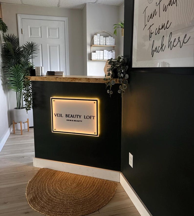 May include: Interior shot of a beauty salon reception area. The black reception desk features a glowing sign that reads "VEIL BEAUTY LOFT SKIN & BEAUTY." A round, woven rug sits on the wooden floor. A framed print with cursive text hangs on the wall.