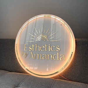 Może przedstawiać: Okrągły, podświetlany szyld z napisem "Esthetics by Amanda" w złotych literach. Szyld ma białe tło, złotą obwódkę i złoty wzór promieni słonecznych.