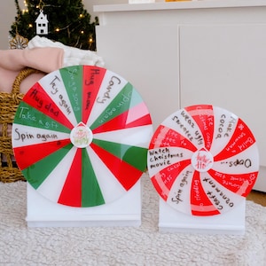 May include: Two Christmas prize wheels in red, green, and white, mounted on white stands. The wheels have handwritten prompts such as "Spin again", "Hug dad", and "Watch Christmas movie". The wheels are a festive decoration.