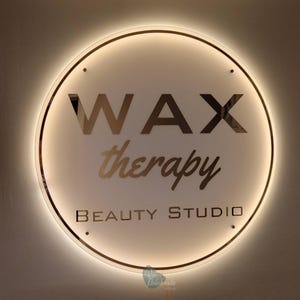 May include: Circular illuminated sign with the words "WAX therapy BEAUTY STUDIO" in gold lettering. The sign has a white background and a gold border, with a warm, inviting glow.