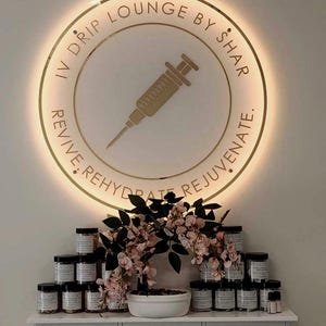 May include: A circular, gold-coloured illuminated sign with the text "IV DRIP LOUNGE BY SHAR" and a syringe graphic. Below, a white shelf displays numerous jars with black lids, alongside a floral arrangement with pink blossoms.