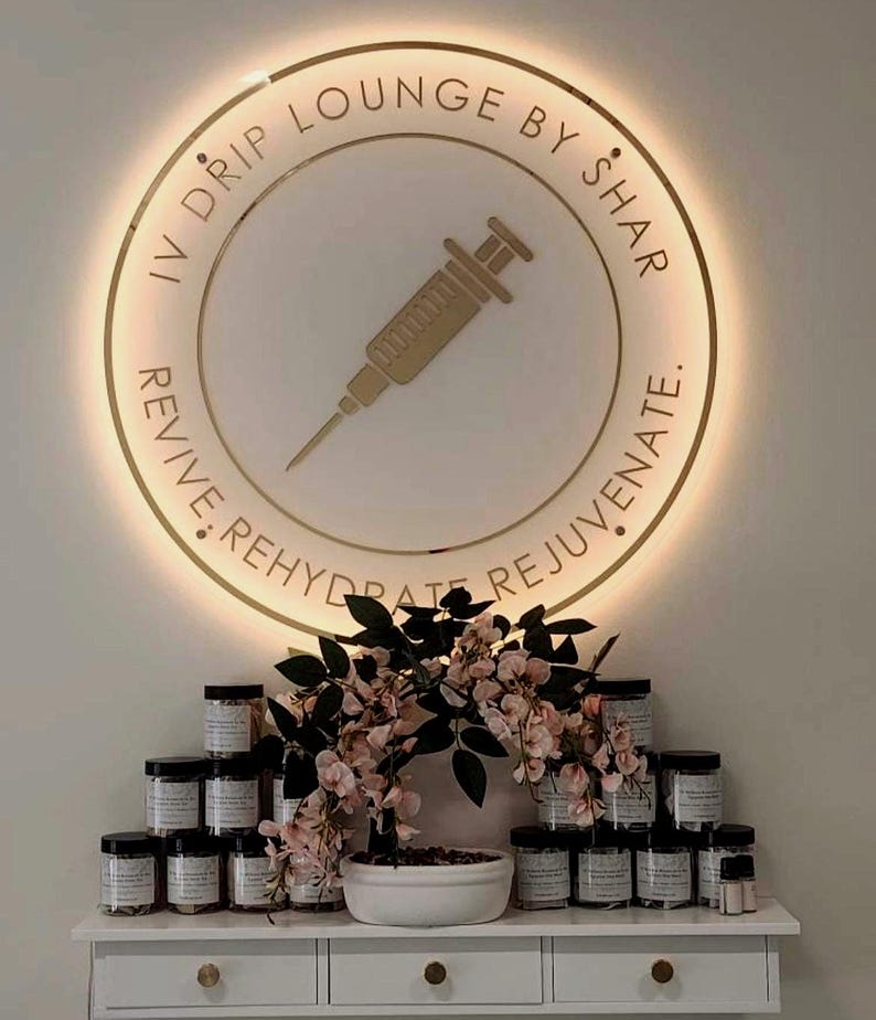May include: A circular, gold-coloured illuminated sign with the text "IV DRIP LOUNGE BY SHAR" and a syringe graphic. Below, a white shelf displays numerous jars with black lids, alongside a floral arrangement with pink blossoms.