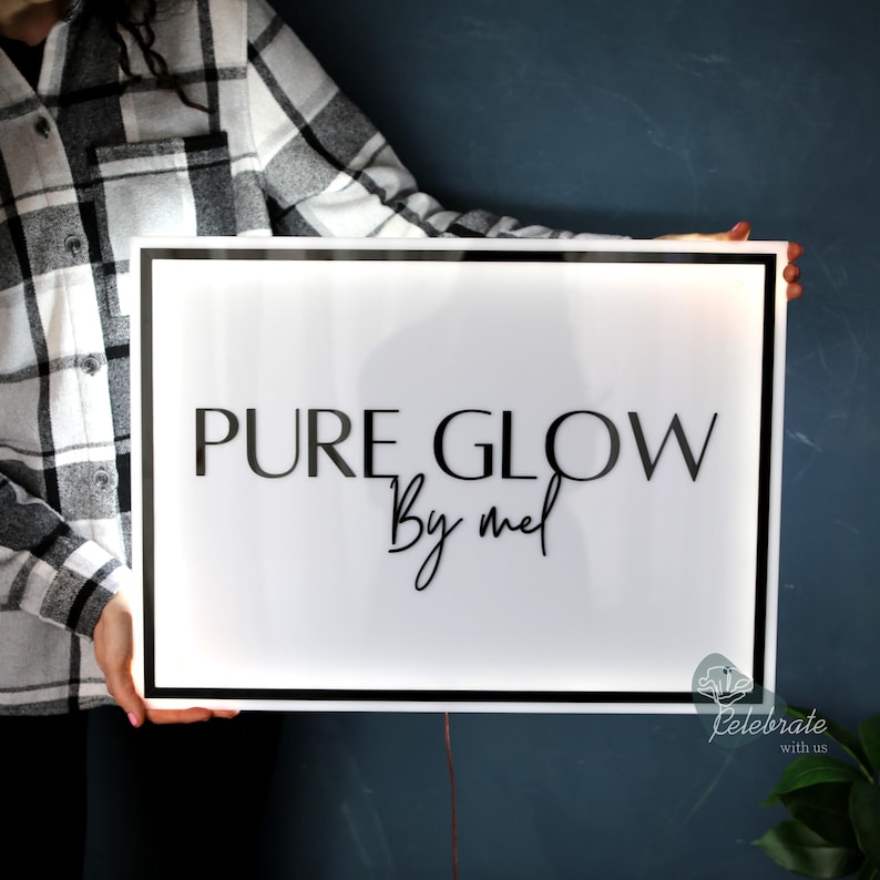May include: A rectangular white sign with the words "PURE GLOW By mel" in black lettering. The sign has a black border and is held up against a dark blue background. The sign is illuminated.
