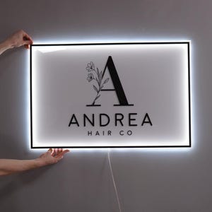 Puede incluir: Un letrero rectangular blanco con un borde negro y letras negras. El letrero tiene un diseño floral negro y la letra "A" con el texto "ANDREA HAIR CO" debajo. El letrero está iluminado con luces LED blancas.