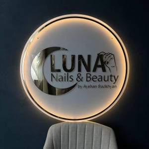 May include: Circular illuminated sign for "LUNA Nails & Beauty" with a crescent moon design. The sign features the business name and a hand illustration. The sign is mounted on a dark blue wall, with a grey chair visible below.