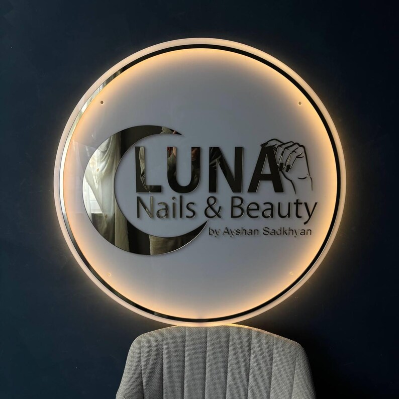 May include: Circular illuminated sign for "LUNA Nails & Beauty" with a crescent moon design. The sign features the business name and a hand illustration. The sign is mounted on a dark blue wall, with a grey chair visible below.