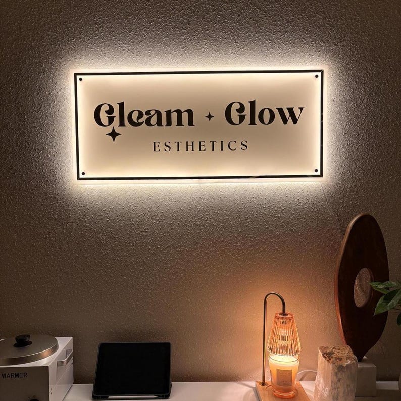 May include: A rectangular illuminated sign with the words "Gleam + Glow ESTHETICS" in black lettering. The sign is mounted on a textured wall. A small lamp and other decorative items are on a white surface below the sign.