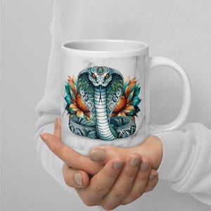 Peut inclure: Une tasse à café en céramique blanche avec un motif marbré. La tasse présente une illustration colorée d'un cobra avec un motif floral.