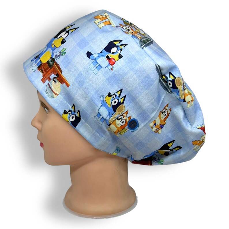 K&ouml;nnte beinhalten: Eine hellblaue OP-Haube mit einem sich wiederholenden Muster von Zeichentrickhunden. Die Haube hat einen karierten Hintergrund und besteht aus einem weichen, flexiblen Stoff. Die Zeichentrickhunde sind in Blau-, Orange- und Schwarzt&ouml;nen gehalten.