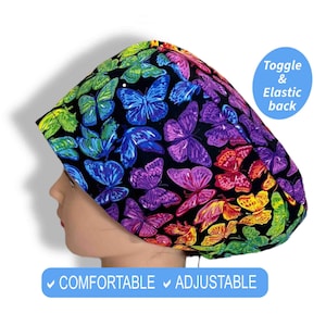 May include: A scrub cap with a rainbow butterfly design on a black background. The cap features a toggle and elastic back. The text "COMFORTABLE" and "ADJUSTABLE" are visible. The cap is designed for comfort and adjustability.
