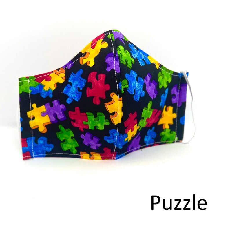 May include: A black face mask with a colorful puzzle piece pattern. The mask has white stitching and a white elastic band.