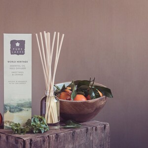 May include: A brown wooden box with a clear glass bottle of essential oil diffuser with wooden sticks. The bottle is labeled "Pure Lakes World Heritage Essential Oil Reed Diffuser Sweet Basil & Orange". There are oranges and green leaves in a wooden bowl on the box.