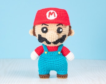 May include: A crocheted Mario doll with a red hat, blue overalls, and a brown mustache. The doll is standing on a white wooden surface.