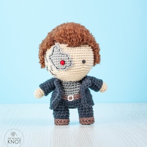 May include: A crocheted amigurumi toy of a character with a red eye, brown hair, and a black jacket. The toy is standing on a light wood surface.