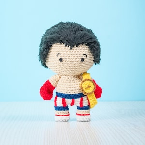 Puede incluir: Un juguete de boxeador de crochet que lleva guantes de boxeo rojos y un cinturón de campeonato amarillo. El juguete tiene el pelo negro y pantalones cortos a rayas blancas y rojas.