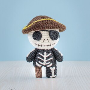 May include: A crocheted skeleton pirate doll wearing a brown hat with a yellow trim and an eye patch. The doll is standing on a light wood surface.