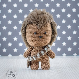 May include: A brown crocheted Chewbacca toy with a white bandolier. The toy is standing on a white wooden surface in front of a gray background with white stars.