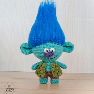 May include: A blue and green crocheted troll doll with a blue fuzzy hairpiece. The troll is wearing a green vest and brown pants.