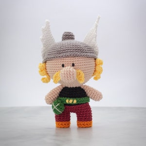Puede incluir: Un juguete de peluche tejido a crochet de un vikingo con un casco gris, cuernos blancos, barba amarilla, nariz marrón, camisa negra, cinturón verde, pantalones rojos y zapatos naranjas.