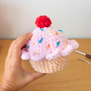 Op de afbeelding: Een handgemaakte gehaakte cupcake met roze glazuur, kleurrijke hagelslag en een rode pomponkers. De cupcake heeft een lichtbruine basis. Haakbenodigdheden zijn op de achtergrond.