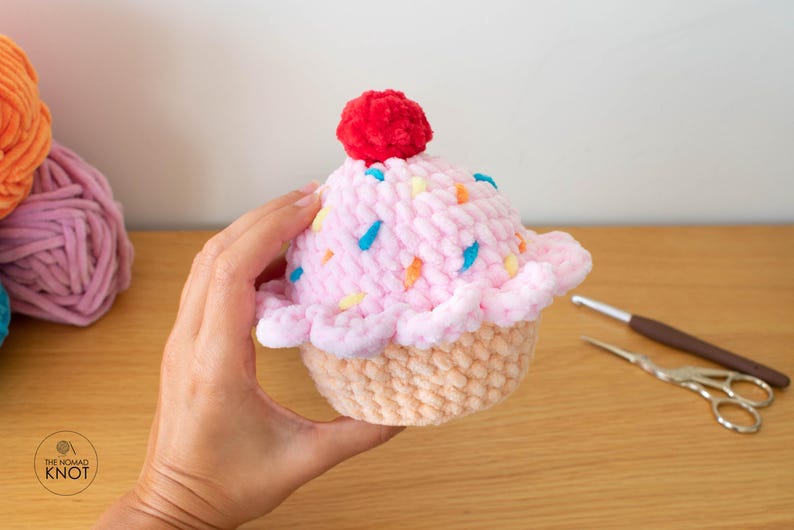 Op de afbeelding: Een handgemaakte gehaakte cupcake met roze glazuur, kleurrijke hagelslag en een rode pomponkers. De cupcake heeft een lichtbruine basis. Haakbenodigdheden zijn op de achtergrond.