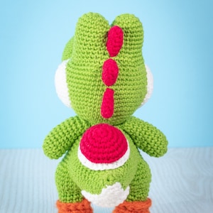 May include: A green crocheted Yoshi plush toy with red and white accents. The toy is standing on a wooden surface.