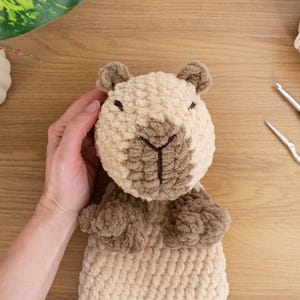 May include: A handmade, plush capybara toy in shades of beige and brown. The crochet animal has small ears, a detailed face, and small paws. A crochet hook and scissors are next to the toy. The Nomad Knot logo is in the bottom left corner.