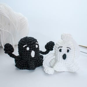 May include: Two crocheted ghosts, one black and one white, with googly eyes and open mouths. The ghosts are standing on a white surface.