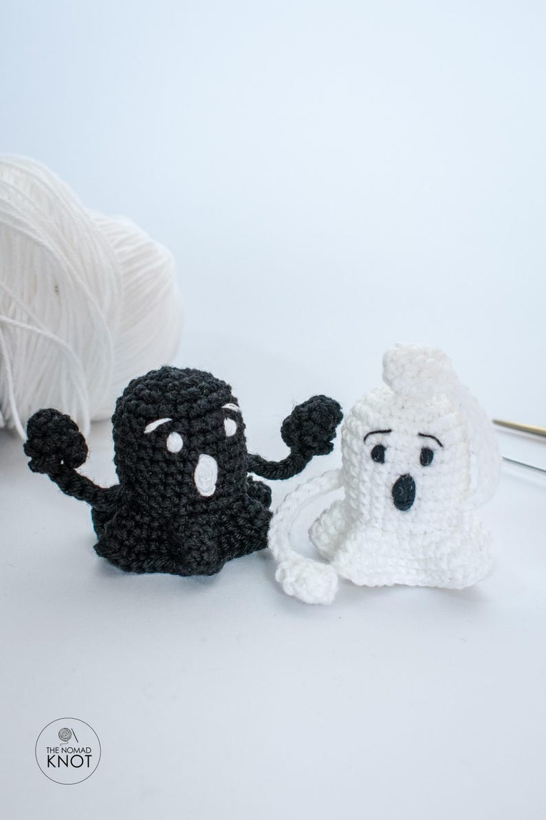 May include: Two crocheted ghosts, one black and one white, with googly eyes and open mouths. The ghosts are standing on a white surface.